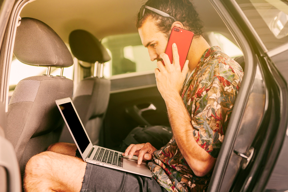 A man in a car looking at his laptop while on the phone.