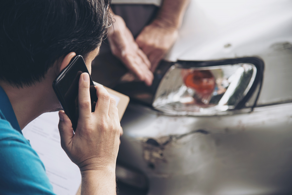 A dark haired man on mobile phone looking at a damaged car.