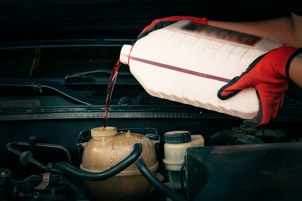 Pair of red-gloved hands pouring antifreeze into a car's engine coolant reservoir.
