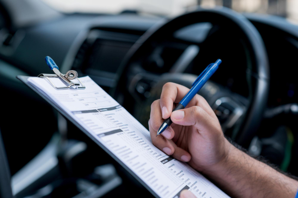 A hand holding a pen above a clipboard in front of a car's steering wheel.